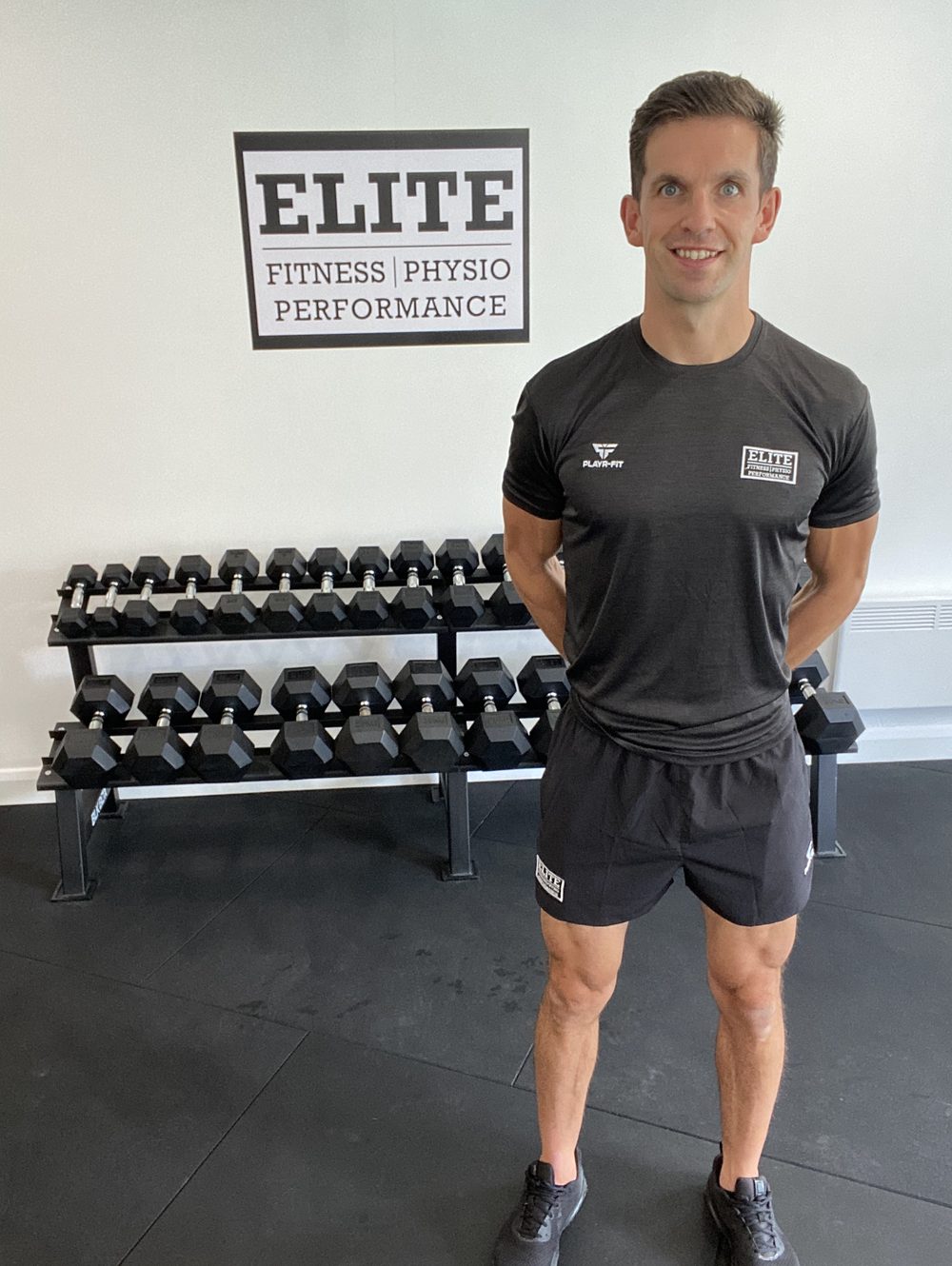 Stephen in Elite kit standing in front of the framed Elite sign and a dumbbell rack
