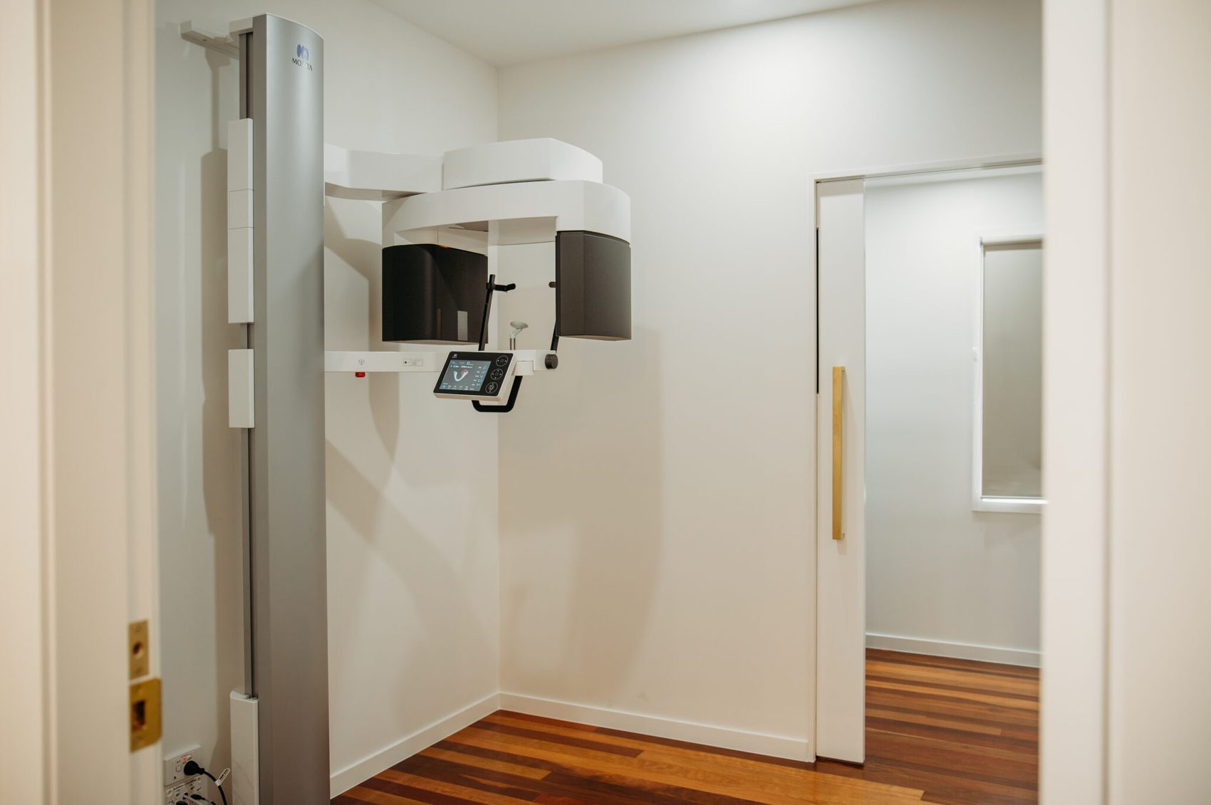 Wall-mounted imaging unit on a column beside an open doorway, with white walls and a timber floored corridor beyond