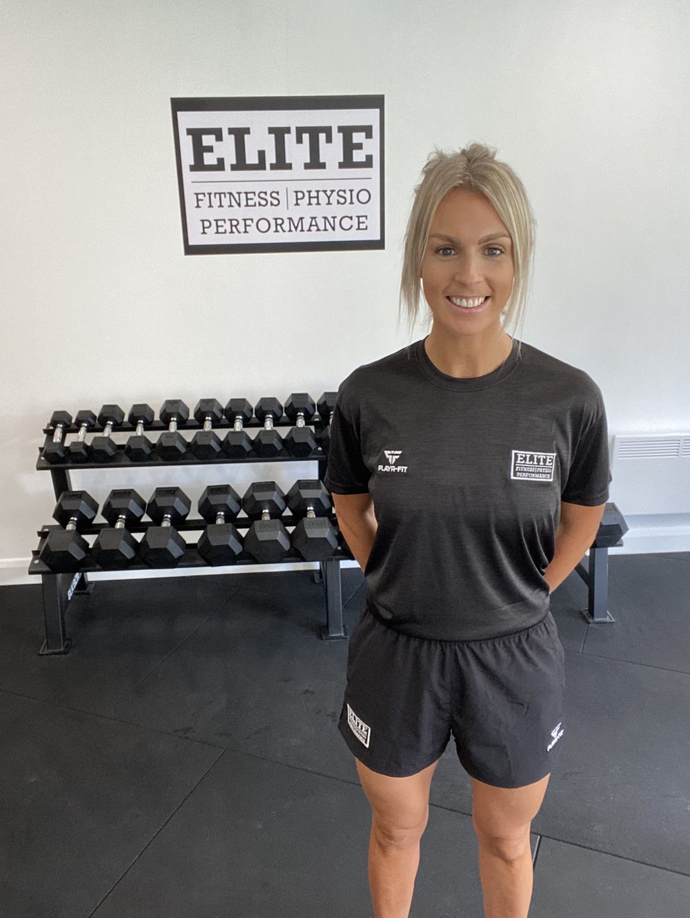Laura in Elite kit standing in front of the framed Elite sign and a dumbbell rack