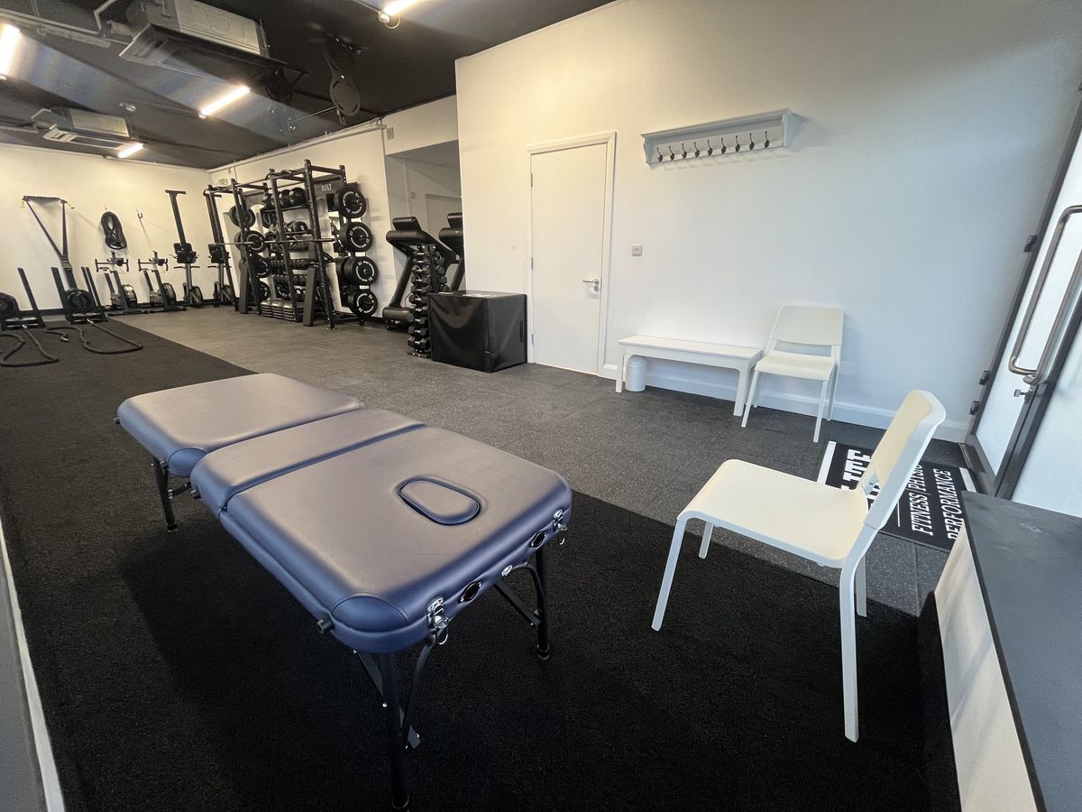 A treatment table and chair in the physio room with the gym racks visible behind