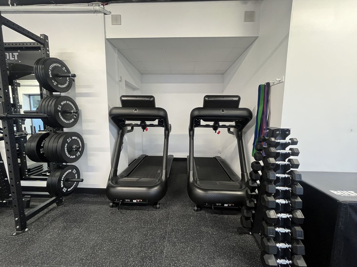 Two treadmills beside wall-mounted weight plates and a dumbbell rack